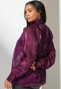 Blusa viola, trasparente, con retro plissettato, polsini aderenti e colletto alto. Tessuto liscio e leggero con una leggera lucentezza.