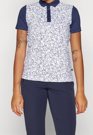 Personne portant un polo bleu marine à manches courtes avec un motif abstrait blanc et bleu marine à l'avant, assorti à un pantalon bleu marine, debout devant un fond uni.
