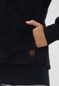 Navy fleece sweatshirt with soft texture, ribbed cuffs, and hem. Features a side pocket and a brown logo patch on the lower hem.