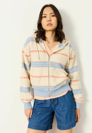 Young woman wearing a cream zip-up hoodie with blue and orange stripes, peach t-shirt, and blue denim shorts, standing against a white background.