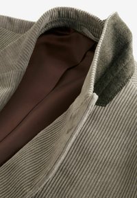 Graue Cordjacke mit einer strukturierten Außenseite, kontrastierender brauner Satin-Innenausstattung und grünen Filzkragenakzenten. Details umfassen vertikale Rippungen.