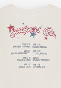 Camiseta beige con texto rojo y azul que dice "Scalpers Co." y varias fechas y ubicaciones impresas en una fuente bold y divertida.
