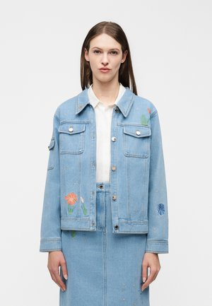 Woman wearing light blue denim jacket and matching skirt with floral embroidery over a white button-up shirt, against plain white background.