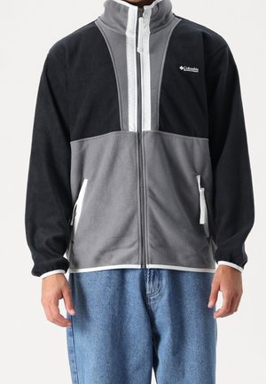 Homme portant une veste polaire Columbia noire et grise avec des fermetures éclair blanches et des accents de poche, associée à un jean bleu.