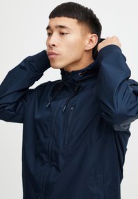 Laivasilkkinen vedenpitävä takki, jossa on vetoketjullinen etuosa, säädettävä huppu ja nyöri. Siinä on rintatasku ja joustavat hihansuut. Sujuva pinta.