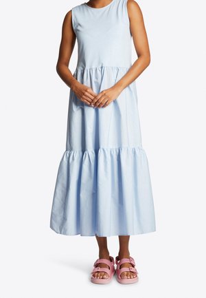 Vrouw draagt een mouwloze lichtblauwe midi-jurk met lagen en roze open sandalen, staat met haar handen voor zich geklemd.