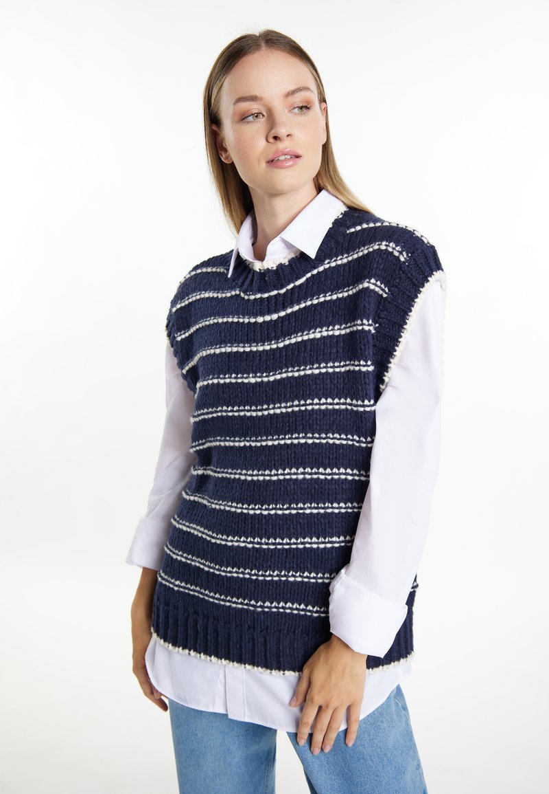 Gilet lavorato a maglia blu navy con strisce orizzontali bianche, orlo a coste e design senza maniche, indossato sopra una lunga camicia bianca.