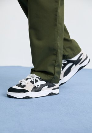 Sorte og hvide sneakers med en tekstureret overdel, der har suede-detaljer og hvide snørebånd, parret med olivengrønne bukser.