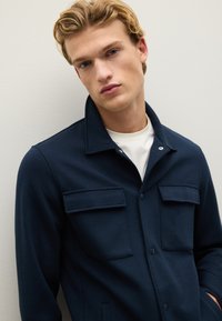 Giacca blu navy con collo, due tasche sul petto e chiusura a bottoni. Realizzata in tessuto liscio e resistente con un design aderente.