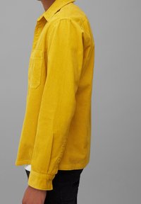 Camisa de pana amarilla con un bolsillo en el pecho, puños con botones y un ajuste relajado. La textura acanalada vertical es distintiva y prominente.