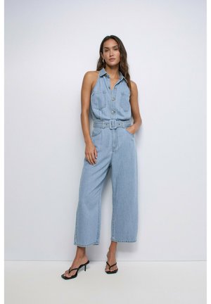 Mujer con mono de mezclilla azul claro sin mangas, pernera ancha, cintura ceñida con cinturón, bolsillos y sandalias de tacón negras, de pie junto a una pared blanca.