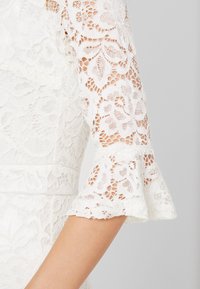Manche de robe en dentelle blanche avec motif floral, tissu semi-transparent et manchette évasée à volants, portée au bras sur un fond uni.