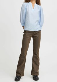 Blouse bleu clair avec des manches bouffantes, décolleté en V et tissu texturé, associée à un pantalon marron évasé et des bottines noires.