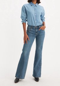Chemise en denim bleu clair à boutons avec manches retroussées, associée à un jean évasé bleu foncé. Chaussures noires pointues complètent le look.