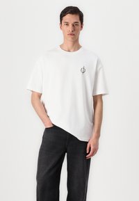 White cotton t-shirt with a round neckline and short sleeves, featuring a black circular logo on the left chest and paired with dark jeans.