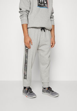 Mann trägt hellgraue Under Armour Jogginghose mit schwarzem Logostreifen an der Seite und graue Sportschuhe mit schwarzen Schnürsenkeln und orangen Akzenten.
