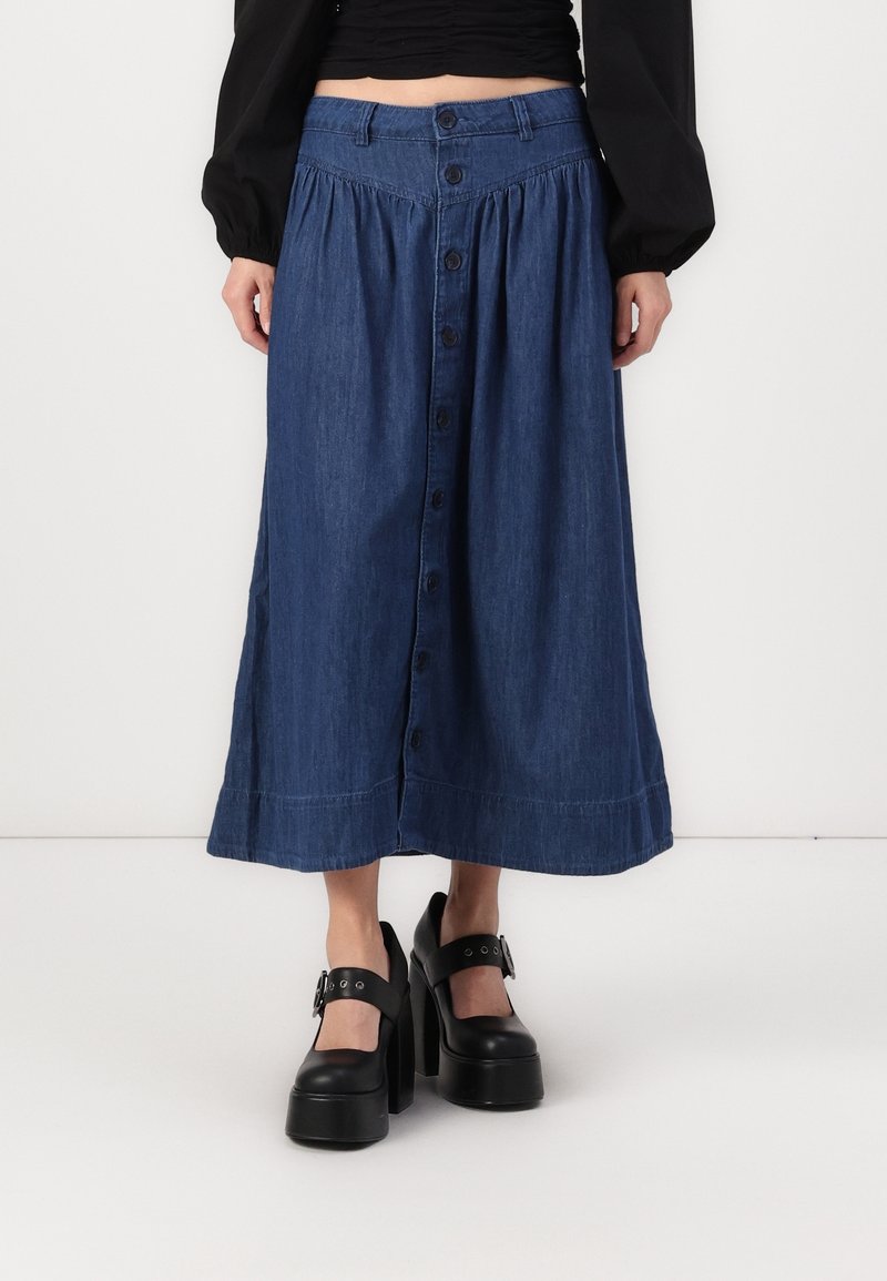 Femme portant une jupe midi en jean bleue taille haute à boutons sur le devant, associée à des chaussures Mary Jane à plateforme noires et un haut noir à manches longues.
