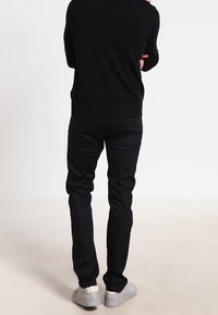 Czarny sweter z prążkowanym wykończeniem, w połączeniu z czarnymi spodniami o wąskim kroju i szarymi sneakersami. Strój jest prosty i elegancki w formie.