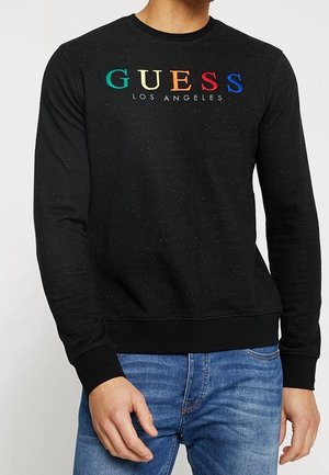Schwarzer Sweatshirt mit langen Ärmeln, verziert mit mehrfarbigen "GUESS"-Buchstaben und dem Schriftzug "LOS ANGELES", kombiniert mit blauen Jeans.