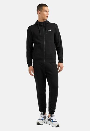 Felpa nera con zip, cappuccio regolabile e logo sul petto, abbinata a pantaloni joggers neri e scarpe da ginnastica bianche.