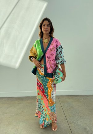 Kvinde står indendørs iført en farverig patchwork-cardigan, matchende lang nederdel, solvedhæng og holder en tekstureret clutchtaske.