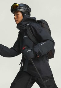 Svart vattentät jacka med texturerat tyg, justerbar huva och logodetaljer. Kombinerad med svarta handskar, en skid hjälm och en ryggsäck.