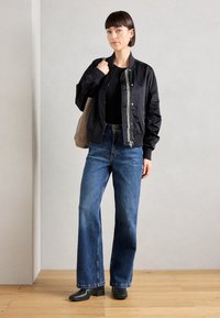 Chaqueta bomber de satén negra con puños acanalados, combinada con jeans de mezclilla anchos de cintura alta en azul y botas negras de tobillo. Bolsa tote en color neutro.