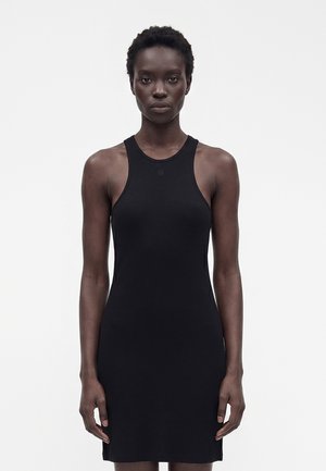 Femme aux cheveux courts et bouclés portant une robe noire moulante sans manches, debout devant un fond blanc uni, regardant droit devant.