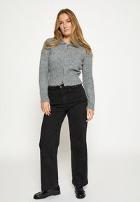 Sivi skraćeni pleteni džemper s patentnim zatvaračem sprijeda i ovratnikom, upareni s crnim hlačama ravnih nogavica i crnim sjajnim cipelama.