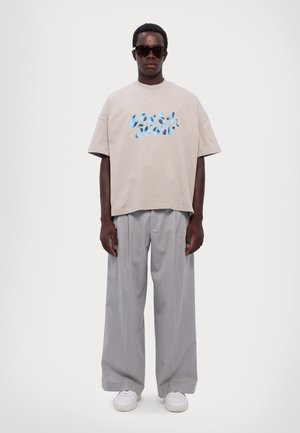 Beżowa bawełniana koszulka z niebieskim grafikiem tekstowym, zestawiona z jasnoszarymi spodniami w kant. Model ma również na sobie czarne okulary przeciwsłoneczne i białe sneakersy.