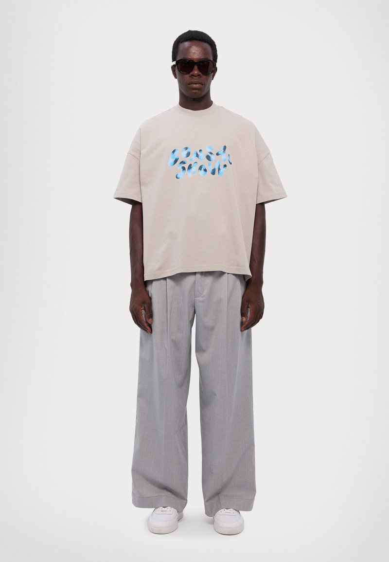 Beige cotton t-shirt with blue graphic text, paired with light gray pleated trousers. Model also wears black sunglasses and white sneakers.