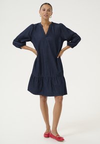 Tmavomodré džínové šaty s výstrihom do V, nariasenými rukávmi a sukňou v poschodiach. Doplnenej červenými plochými topánkami. Jednoduchý, neformálny dizajn.