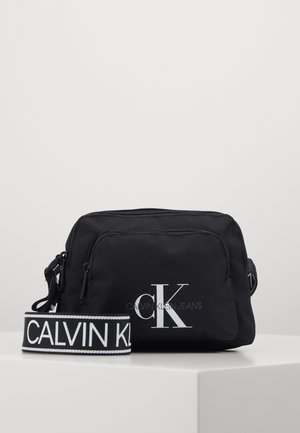 Sac bandoulière noir Calvin Klein avec logo blanc "cK" sur la poche avant et large sangle de marque affichant "Calvin Klein" en texte blanc.