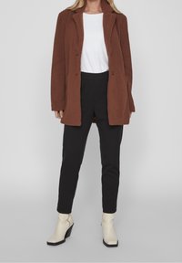 Brun fleecefrakke med reverskrave, sorte bukser og hvid top. Frakken har en tekstureret overflade og to forlommer.