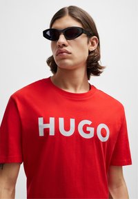 Camiseta de algodón roja con letras blancas "HUGO", combinada con gafas de sol negras elegantes. Diseño de mangas cortas y cuello redondo.