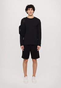 Camisa negra de manga larga y pantalones cortos negros, combinados con zapatillas blancas. Un pequeño bolso cruzado negro descansa sobre el hombro izquierdo.