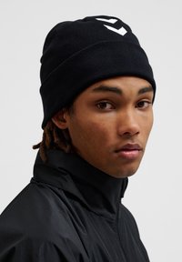 Zwarte gebreide beanie met een ribbelstructuur en een wit logo dat prominent aan de voorkant staat. De muts heeft een comfortabele pasvorm en is zacht.