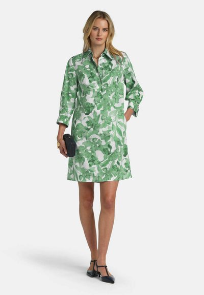 Frau trägt ein weißes knielanges Kleid mit grünem Blumenmuster, hält eine schwarze Clutch und trägt schwarze T-Riemchen-Sandaletten.