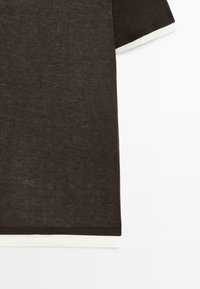 Dunkelbraunes T-Shirt mit kurzen Ärmeln, darüber getragen ein weißer Stoff, sichtbar an Ärmeln und Saum, vor einem schlichten hellen Hintergrund.