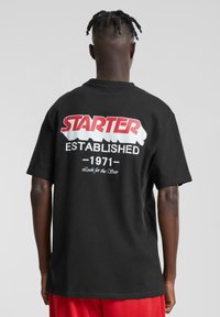 Juodas medvilninis marškinėliai su raudonu ir baltu "STARTER" logotipu ir tekstu "ĮKURTA 1971" spausdintu ant nugaros. Trumpomis rankovėmis ir laisvu siluetu.