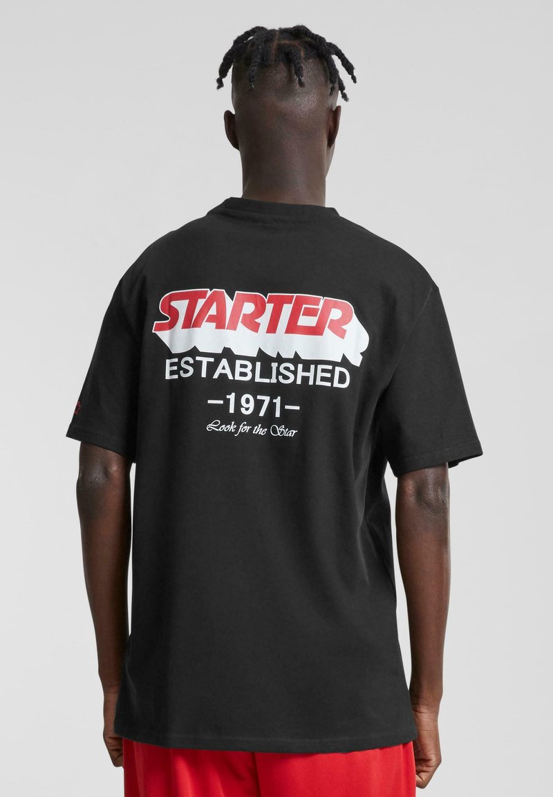 Juodas medvilninis marškinėliai su raudonu ir baltu "STARTER" logotipu ir tekstu "ĮKURTA 1971" spausdintu ant nugaros. Trumpomis rankovėmis ir laisvu siluetu.