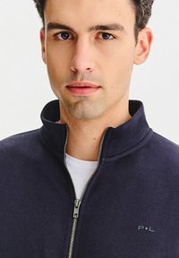 Granatowa kurtka na zamek błyskawiczny z wysokim kołnierzem, wyposażona w srebrny zamek oraz subtelne logo po lewej stronie piersi. Miękka w dotyku, zaprojektowana z myślą o komforcie.