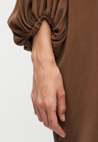 Χέρι που ξεκουράζεται χαλαρά στο πλάι, φορώντας καφέ μακρυμάνικο ένδυμα με ελαστικό μανσέτα που συγκεντρώνει υλικό δημιουργώντας ρυτιδωτή υφή.