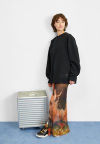 Sudadera negra de gran tamaño, falda larga con un patrón colorido, caja metálica plateada sobre un suelo azul, persona que lleva zapatillas negras y blancas.