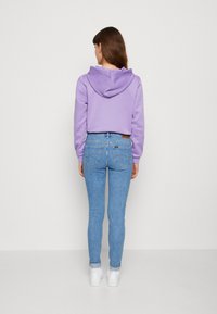 Haut cropped violet avec une grande capuche associé à un jean skinny bleu. La tenue est complétée par des baskets blanches et un design simple.