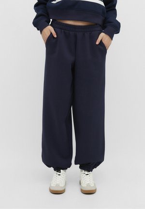 Personne portant un pantalon de survêtement large bleu marine avec des poignets élastiques et des baskets blanches, les mains dans les poches, debout devant un fond uni.