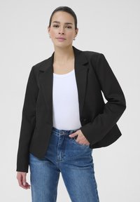 Schwarzes Blazer mit einem einzigen Knopfverschluss, schmal zulaufenden Revers und taillierter Passform, über einem weißen Oberteil getragen und mit hochgeschnittenen blauen Jeans kombiniert.