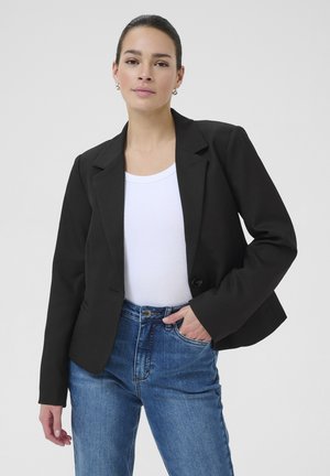 Černý blazer s jedním knoflíkem, klopovými límci a přizpůsobeným střihem, vrstvený přes bílé tričko a kombinovaný s modrými džínami s vysokým pasem.