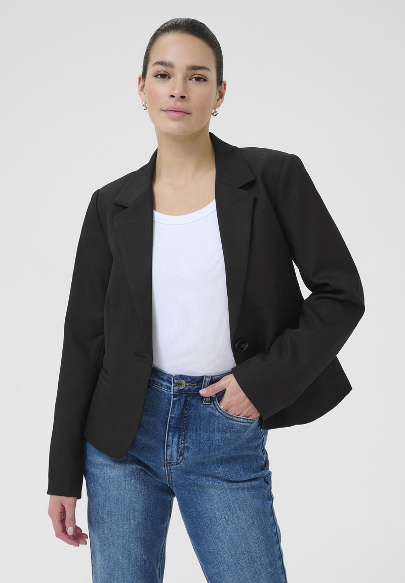 Schwarzes Blazer mit einem einzigen Knopfverschluss, schmal zulaufenden Revers und taillierter Passform, über einem weißen Oberteil getragen und mit hochgeschnittenen blauen Jeans kombiniert.
