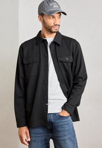 Mand iført sort skjorte over grå T-shirt, blå jeans og grå kasket, står med den ene hånd i lommen og ser sidelæns.
